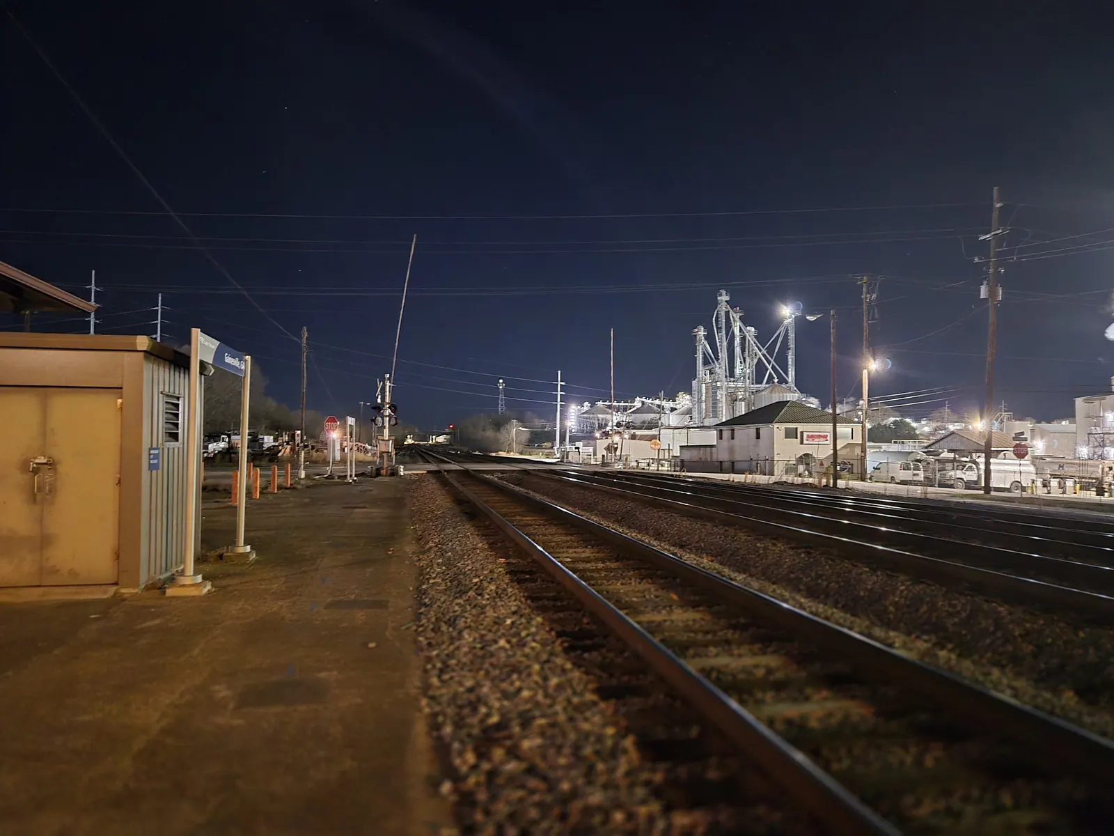 Night view along railroad tracks from a station platform with a small metal equipment shelter and a blue station sign, toward a brightly lit industrial plant with tall towers and a crossing signal.