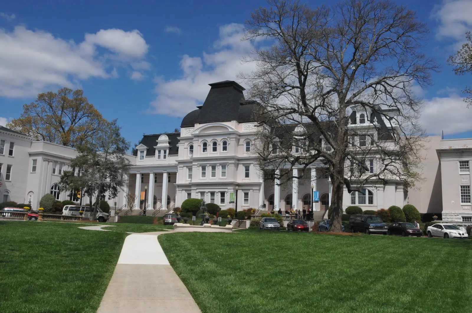 A walkway leads across a green lawn toward a white multi-story building with a dark mansard-roofed central tower and columned entrance, flanked by other white buildings, with bare and leafy trees and parked cars under a blue sky with clouds.