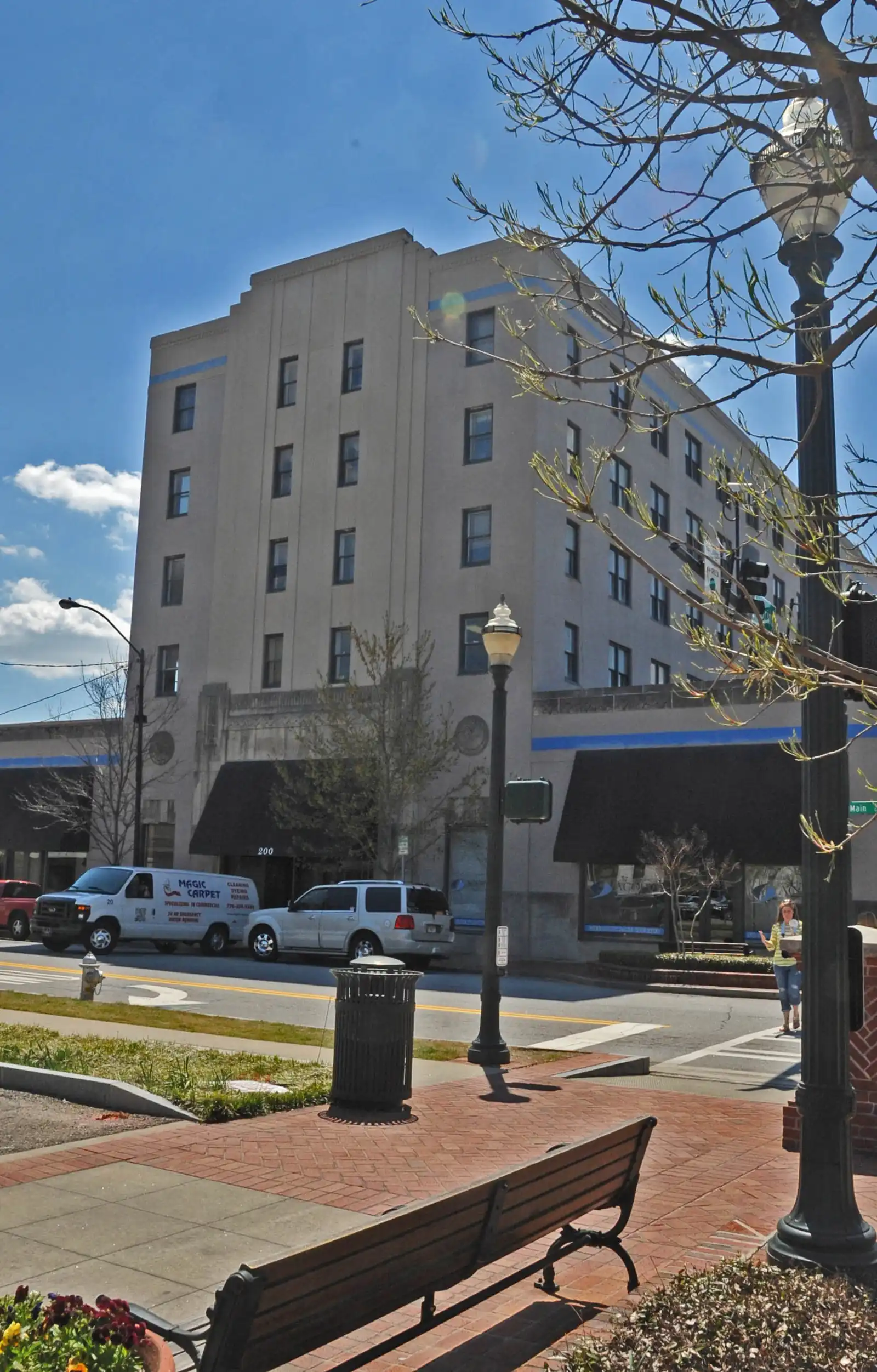 A tall pale Art Deco-style building rises behind a brick plaza with a bench, black lamp posts and a trash can, beside a street with parked vans and bare-branched trees under a blue sky; a pedestrian is visible in the distance.