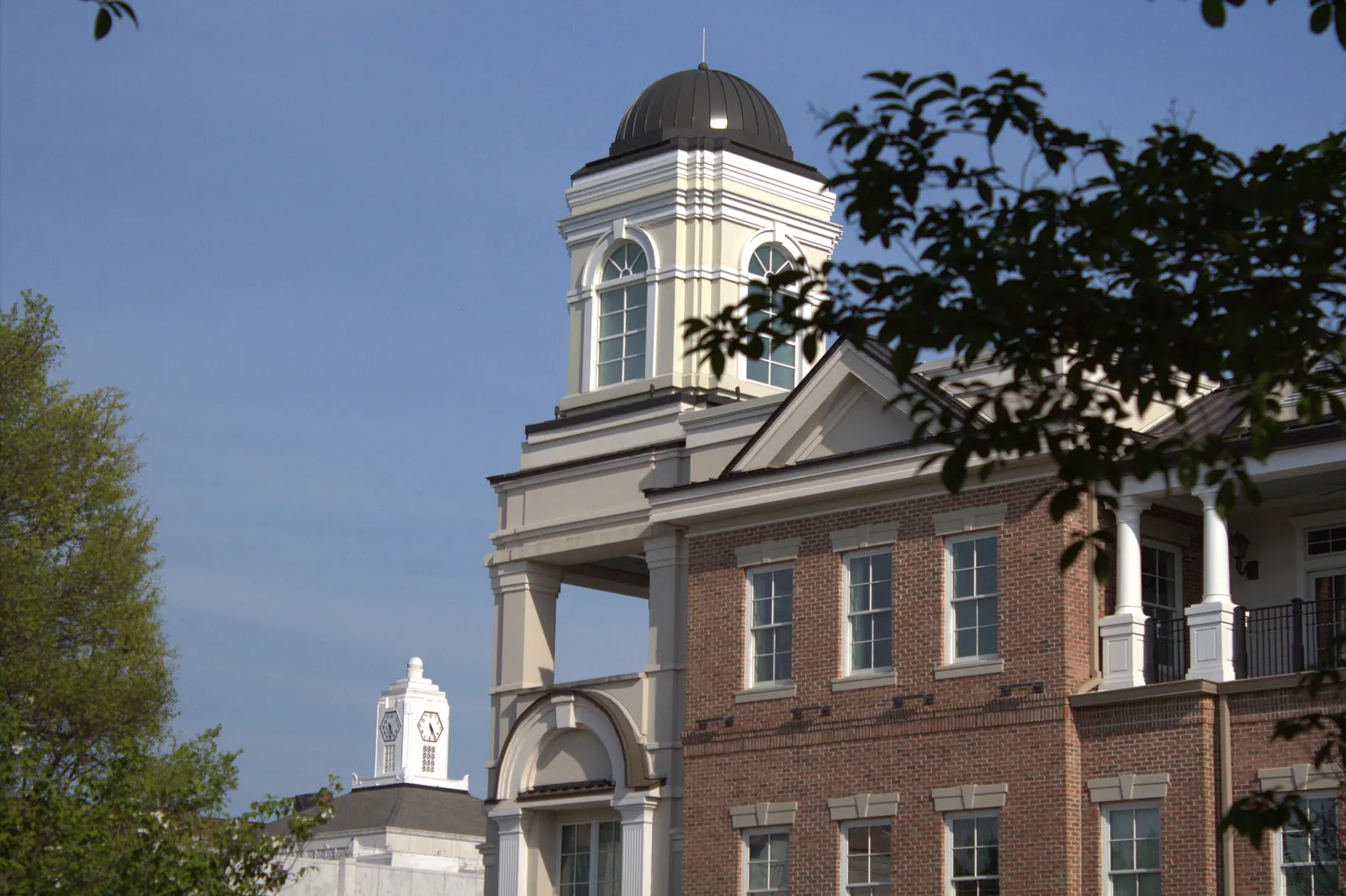 A red-brick building with white columns, an iron-railed balcony and a cream cupola topped by a dark metal dome stands against a blue sky, framed by tree leaves, with a smaller white clock tower visible in the lower-left distance.