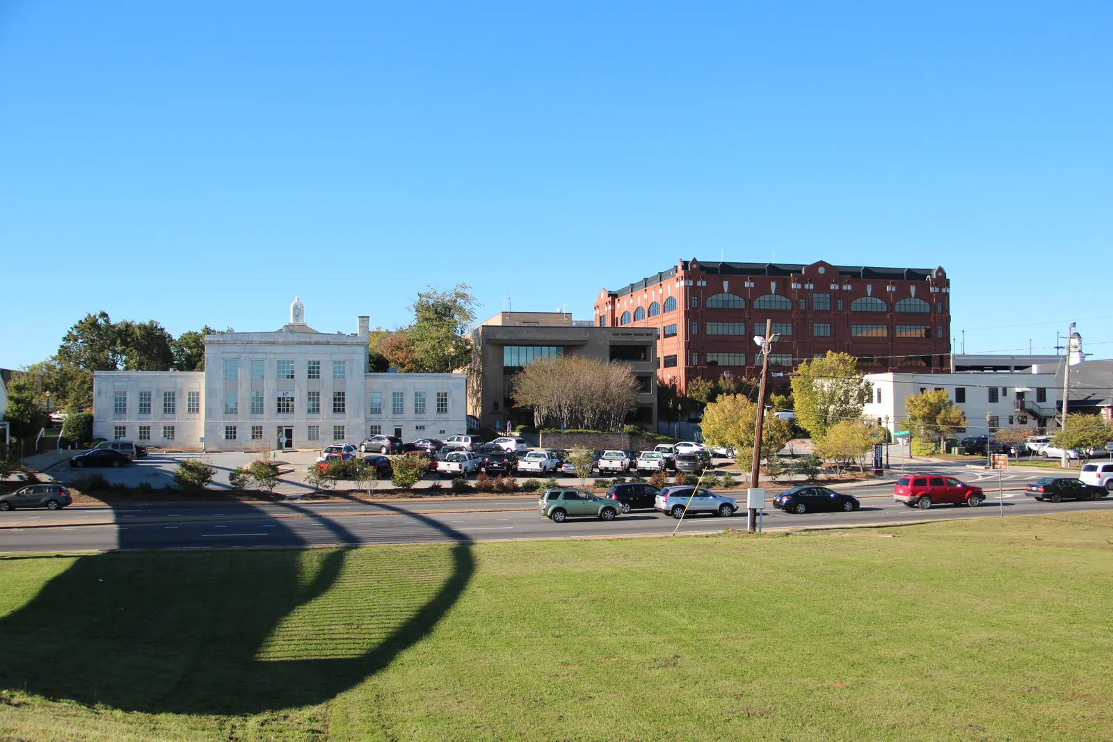 Across a mown lawn and a multi-lane road, a white stone building with a small cupola, a modern office building and a large red-brick building with arched upper windows stand behind a full parking lot under a clear blue sky, with the shadow of a bridge arcing across the grass.
