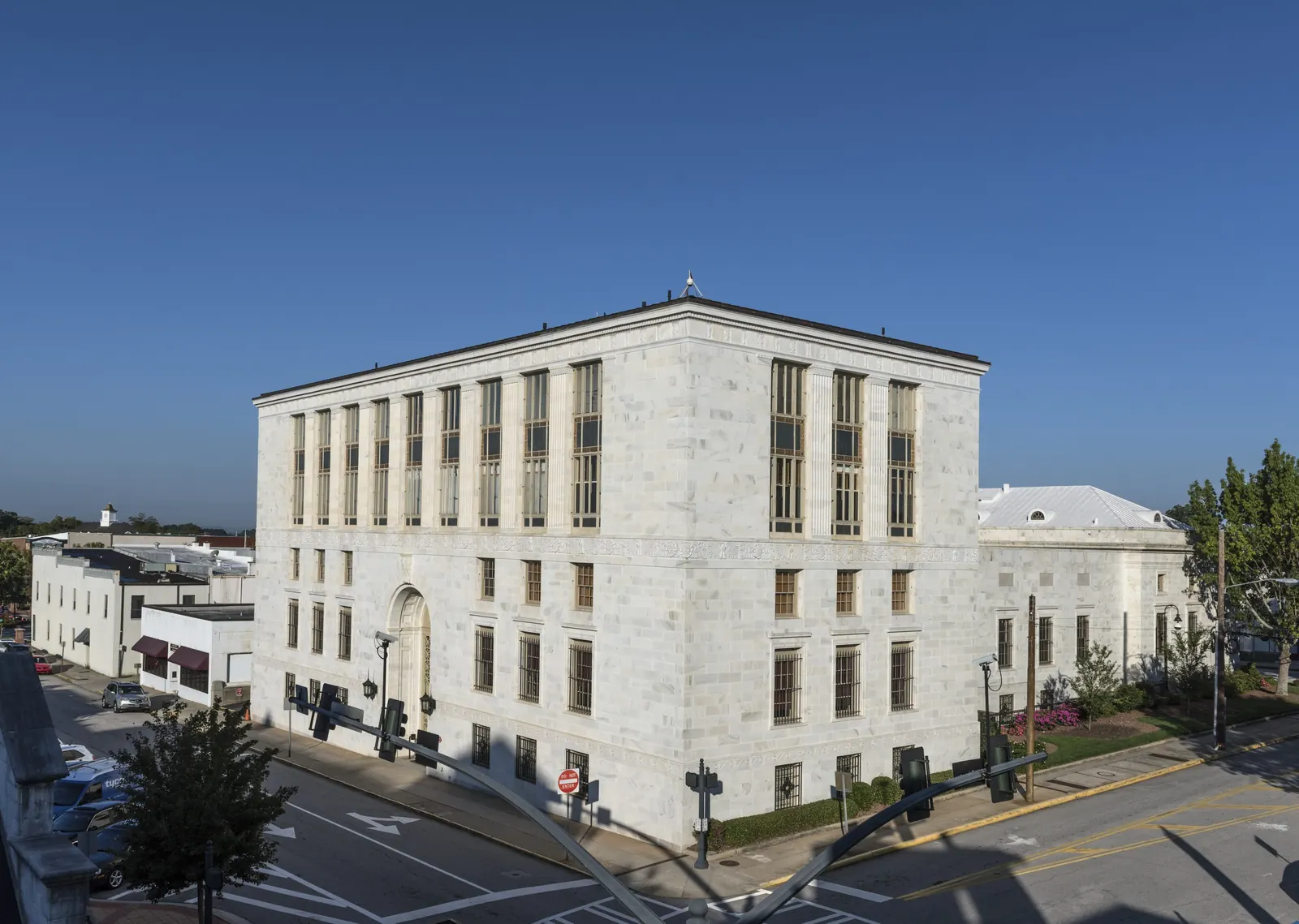 Elevated morning view of a white marble multi-story building on a street corner, with tall vertical window bays, traffic signals in the foreground and low buildings beyond under a clear deep-blue sky.