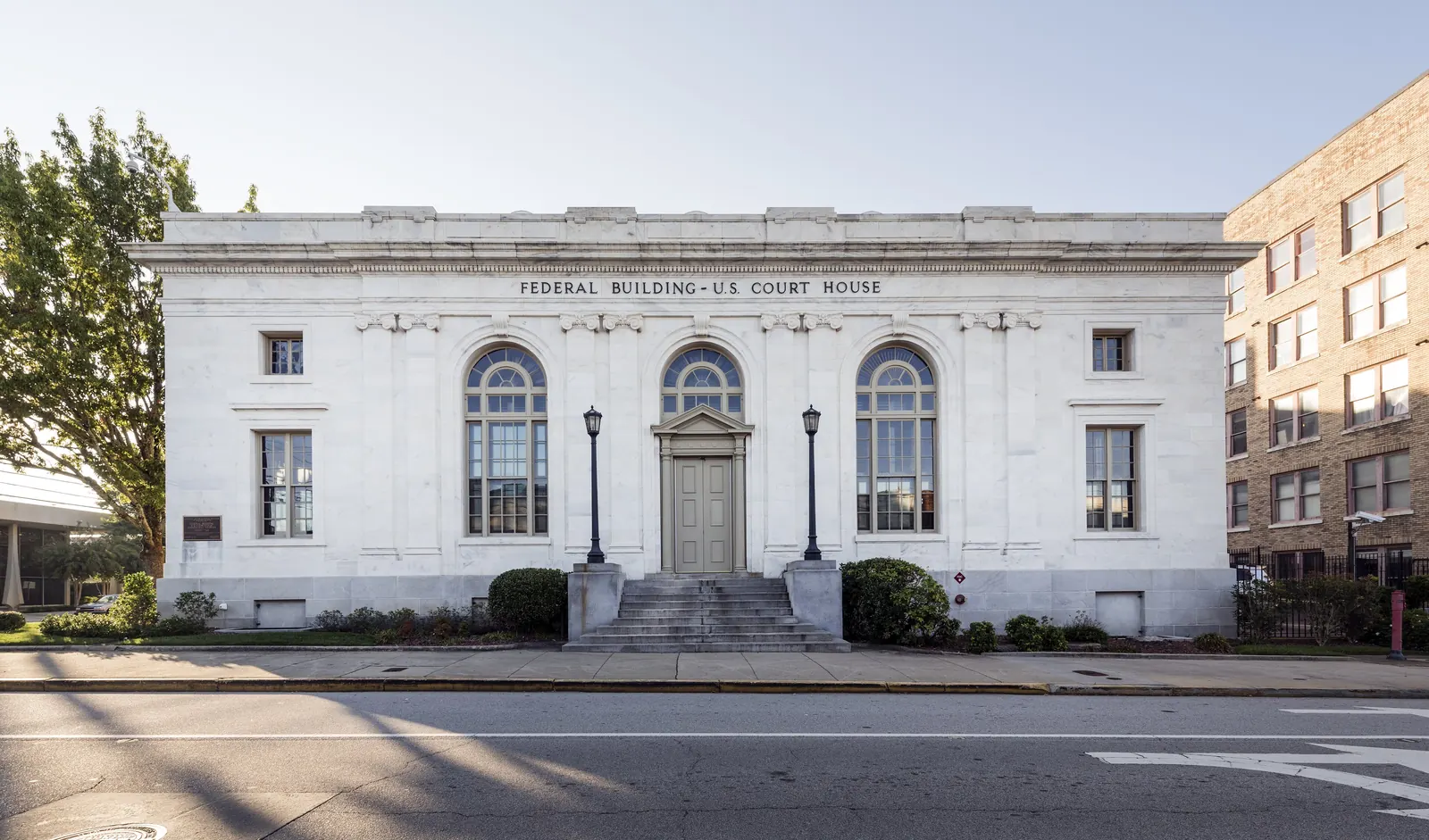 Front of a white marble building with three tall arched windows, a central door up a short flight of steps flanked by lamp posts, and 'FEDERAL BUILDING - U.S. COURT HOUSE' carved above, with a tan brick building at right.