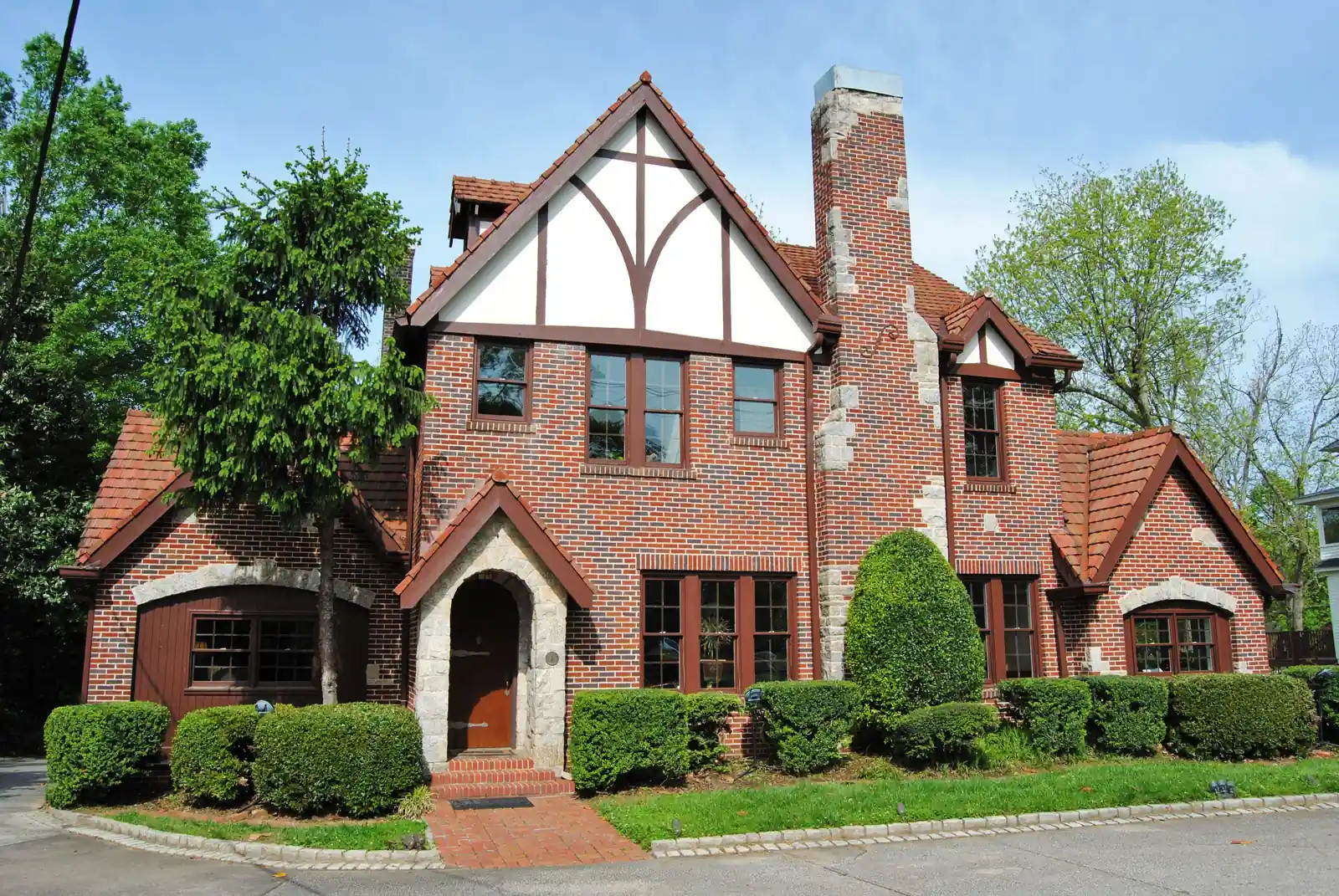 A red-brick Tudor Revival-style building with steep tiled gables, a half-timbered upper gable, a tall chimney and a stone-trimmed arched doorway, fronted by clipped hedges under a blue sky.