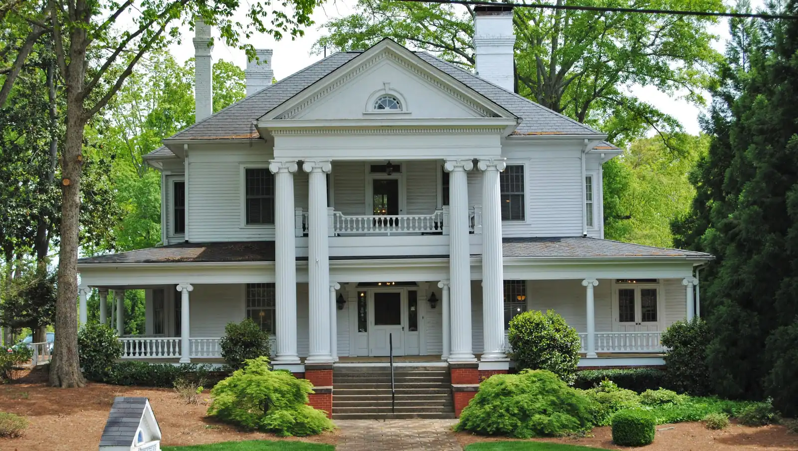 A two-story white wooden house with a tall columned portico, a pediment with a fan window, a wraparound porch and brick chimneys, surrounded by mature trees and shrubs, with a small sign in the front bed.