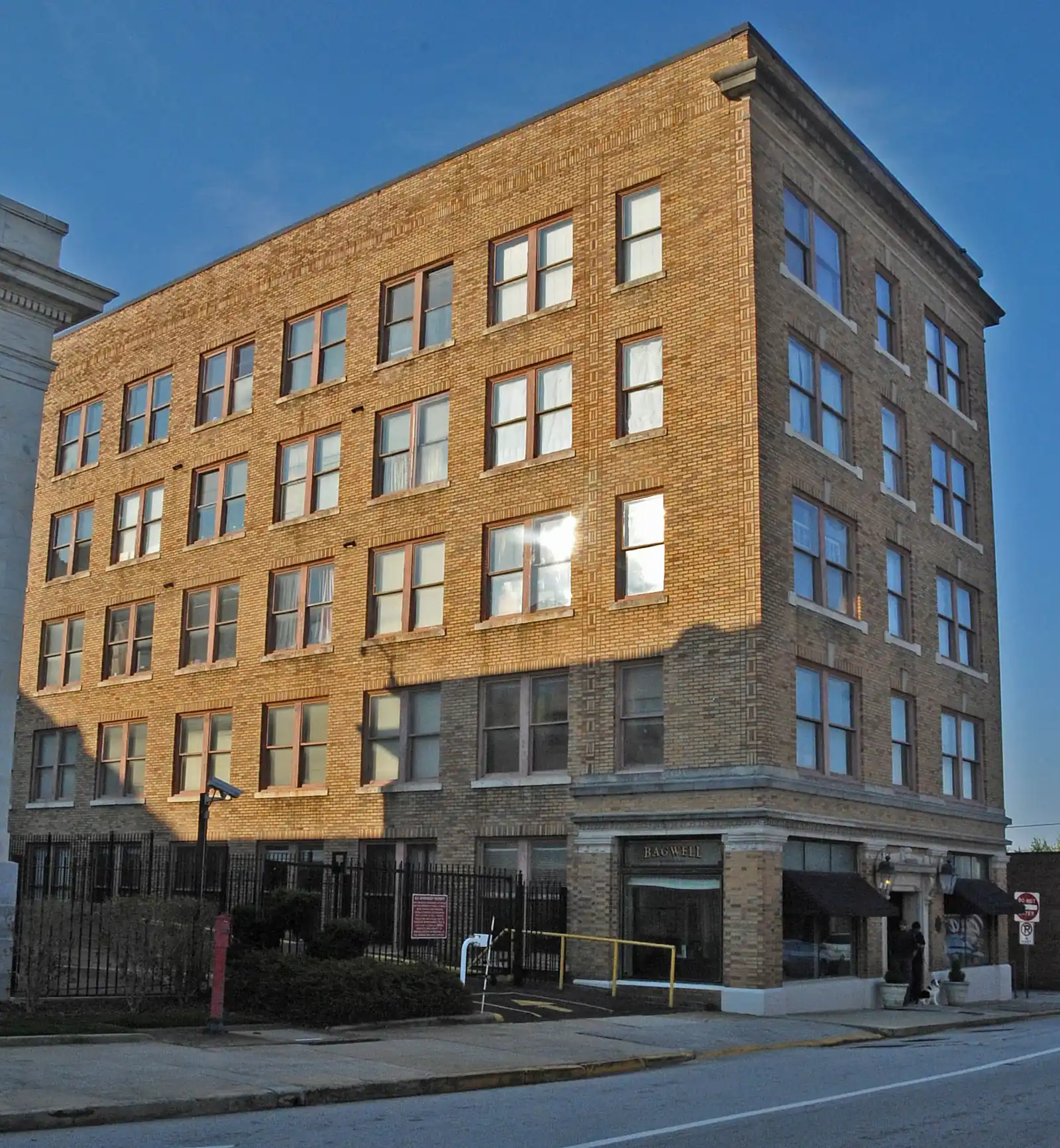 A five-story tan brick building with rows of windows stands on a street corner in strong sunlight, with a black iron fence at its base and the edge of a white stone building at left, under a clear blue sky.