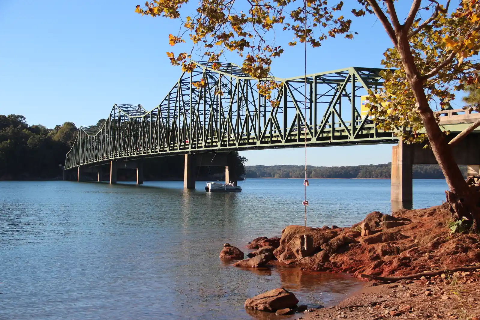 A long green steel truss bridge on concrete piers spans a blue lake, with a pontoon boat beneath it, red rocks along the shore and a tree with yellowing leaves and a hanging rope in the foreground under a clear blue sky.