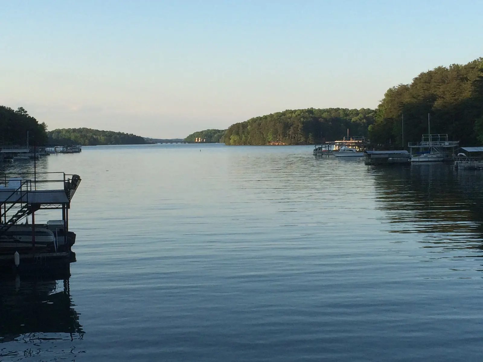 A calm lake cove in soft evening light, lined on both sides by wooded shores with boat docks and moored boats, opening to a distant channel.