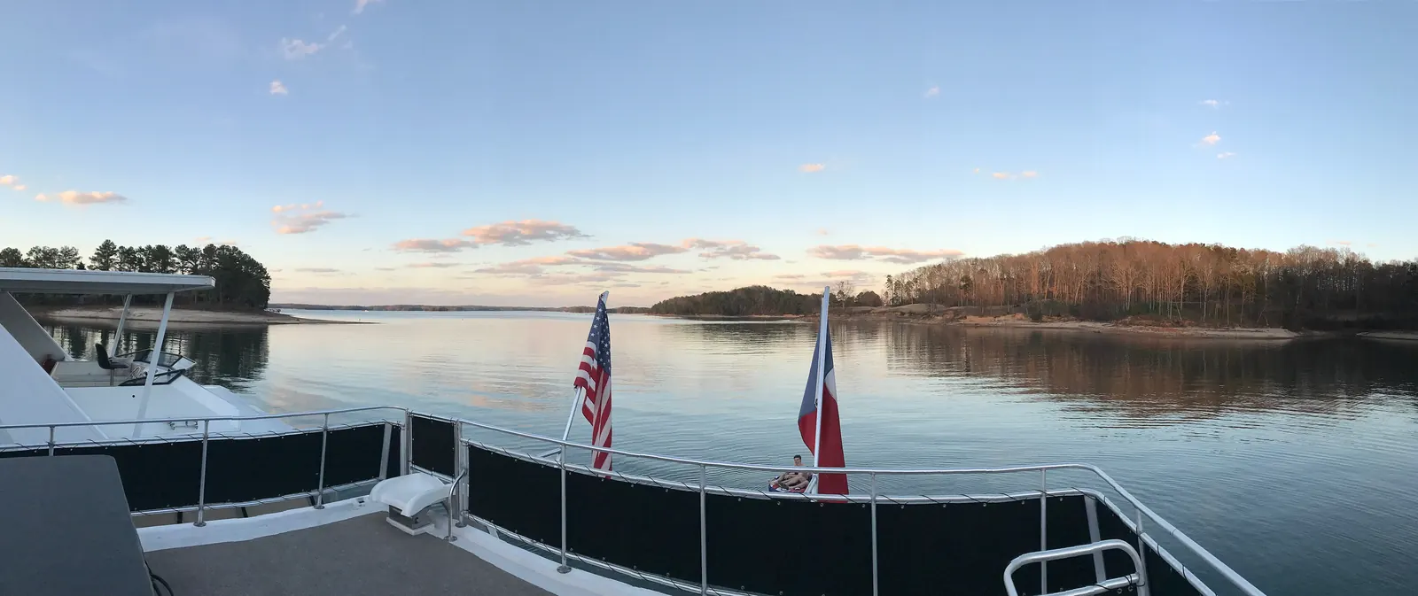 Smartphone panorama from the rear deck of a houseboat across calm water at sunset, with an American flag and a second red, white and blue flag on the railing, wooded shores and pink-tinted clouds; a person sits at the stern in the distance.
