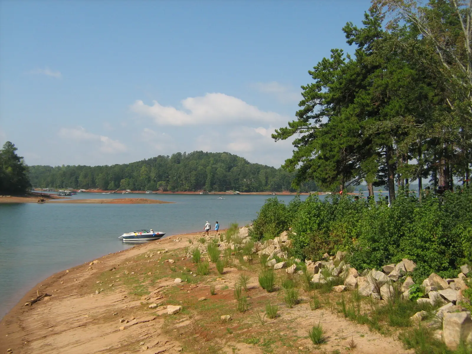 A sandy, rocky shoreline with exposed red clay runs along a lake at low water, with a small motorboat pulled up near two distant people, wooded hills across the water and tall pines at right under a blue sky.