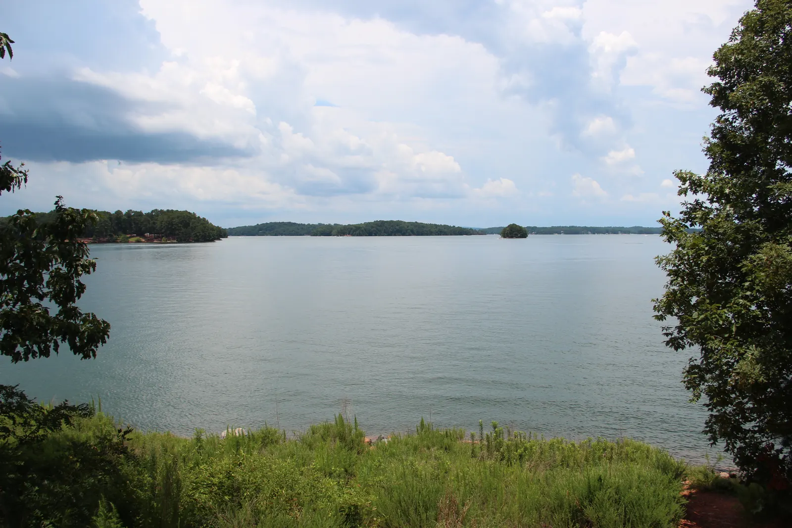 A broad calm lake under billowing white clouds, seen from a grassy bank between trees, with wooded islands and a distant shoreline.