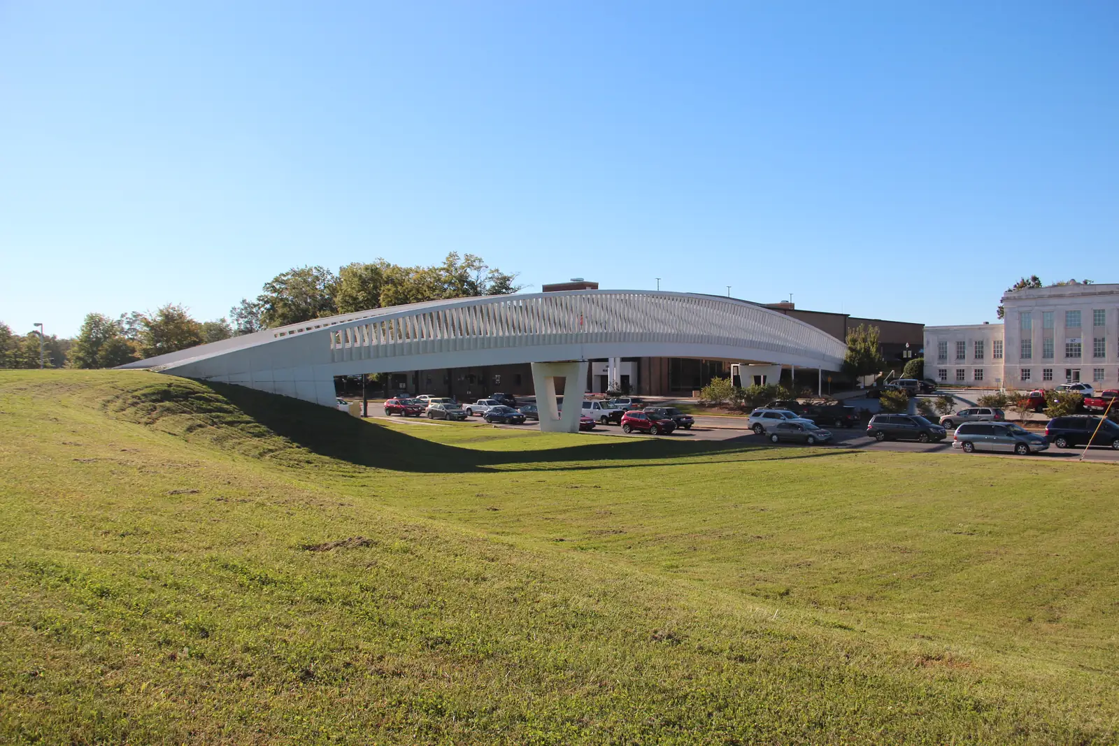 A long white pedestrian bridge with a ribbed canopy arcs over a road and parking lot beyond a grassy slope, with a white stone building at right and trees behind under a blue sky.