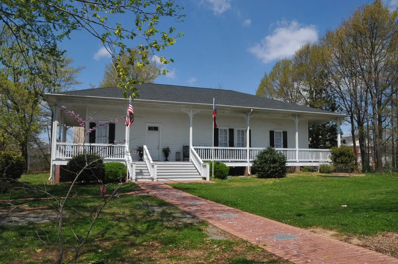 A white one-story clapboard building with a long front porch, black shutters, an American flag and a Georgia state flag stands on a green lawn with a brick walkway, framed by early-spring trees under a blue sky with clouds.