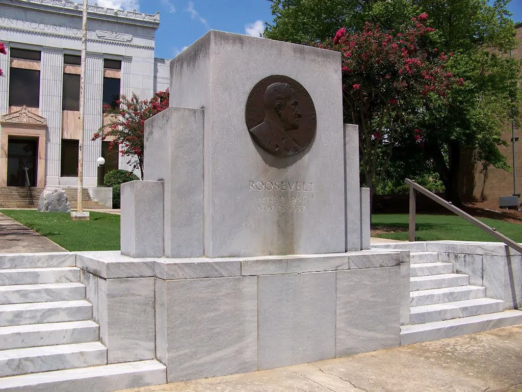 A white marble memorial with a bronze relief portrait medallion above the word 'ROOSEVELT' sits on a stepped marble base in front of a white stone government building with flowering crape myrtles.